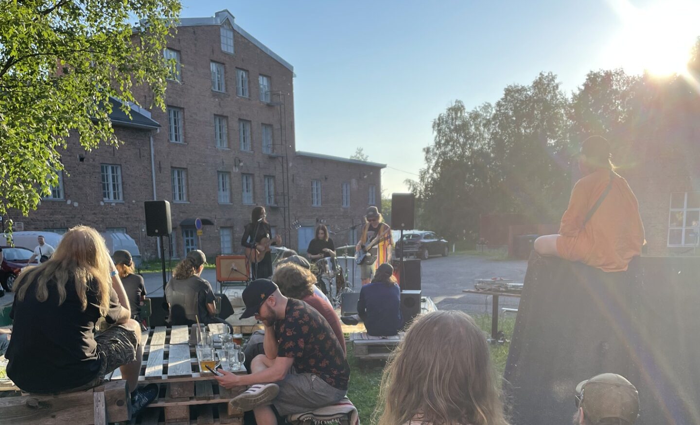 Mallassauna-tapahtuma. Ihmisiä ulkona istuskelemassa, yhtye soittaa musiikkia, kesä, taustalla tiilirakennus.